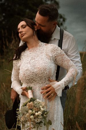 A bride in lace gown holds a bouquet while her groom in suspenders kisses her cheek in a rustic outdoor setting.