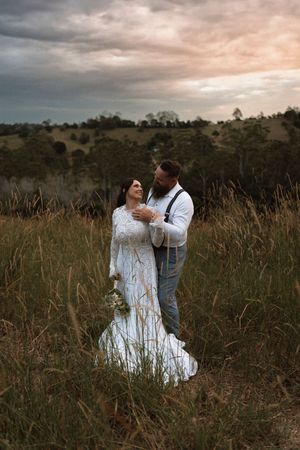 Bride and groom embrace in tall golden grass field at sunset with dramatic cloudy sky and rolling hills backdrop.