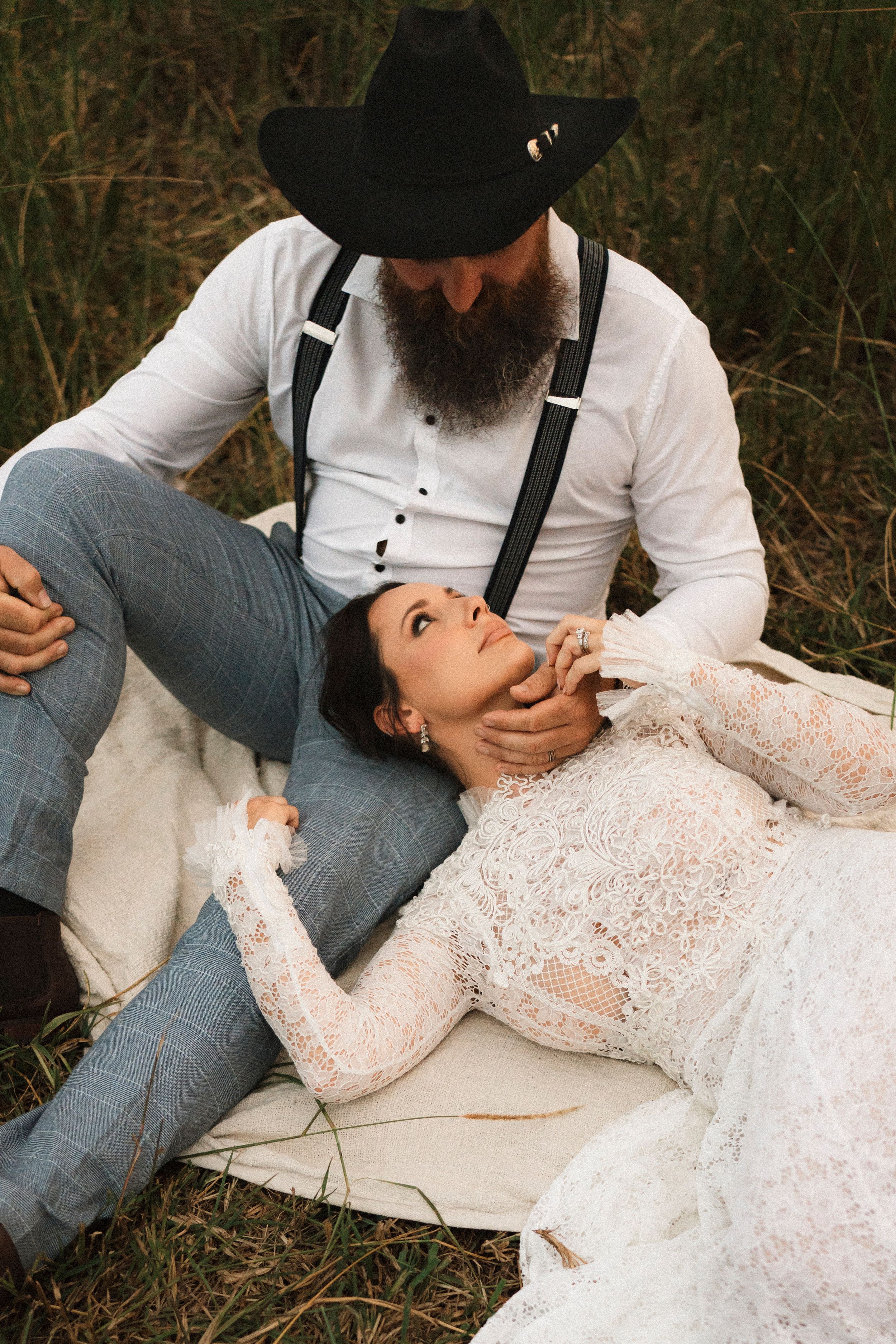 Bearded groom in black cowboy hat and suspenders holds brides face as she lays in his lap outdoors.