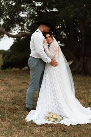 Bride in lace gown with veil embraces groom in cowboy hat outdoors under large tree on wedding day.