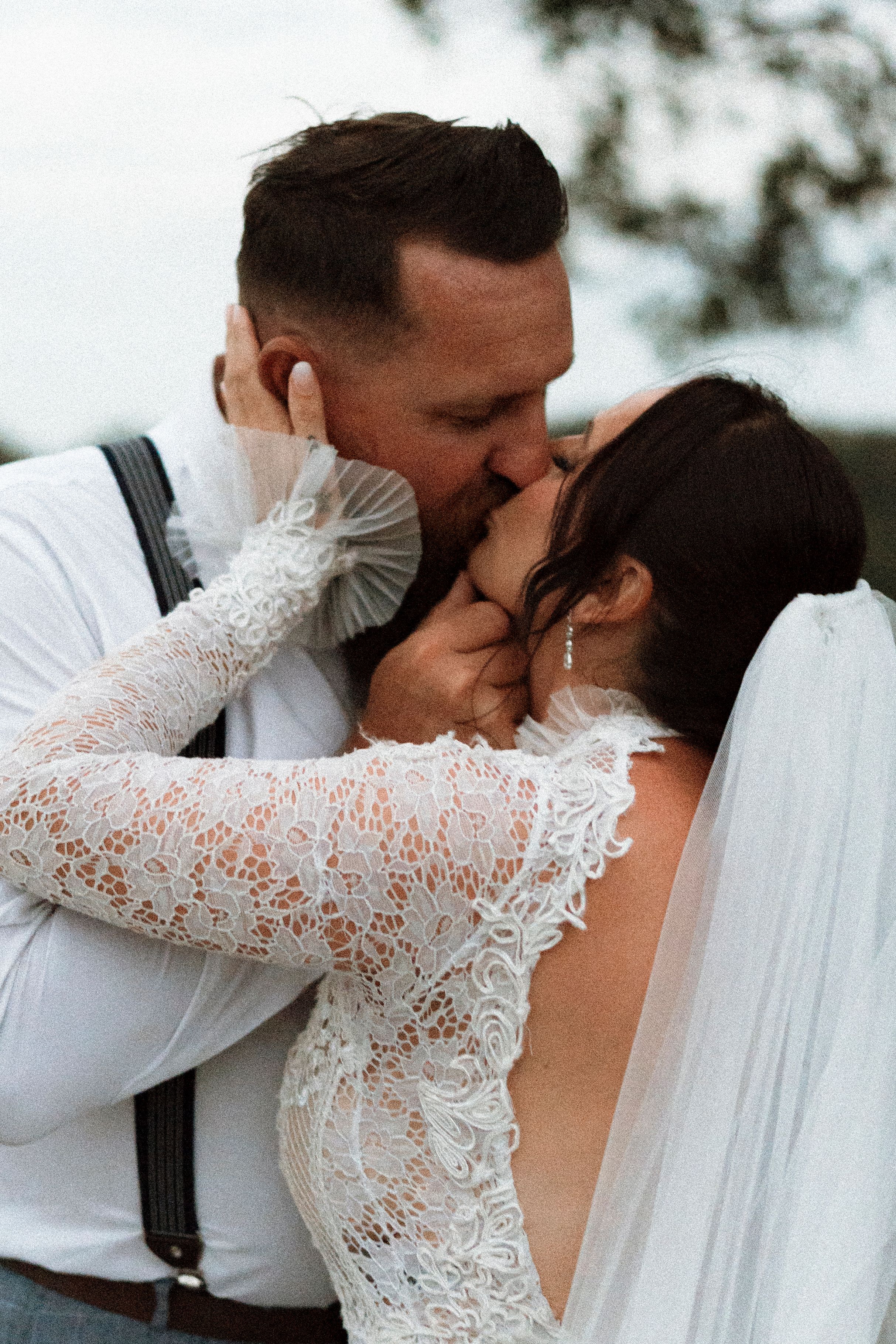 A bride and groom share a romantic first kiss, she wearing a lace long-sleeve wedding gown with veil, he in white shirt with suspenders.