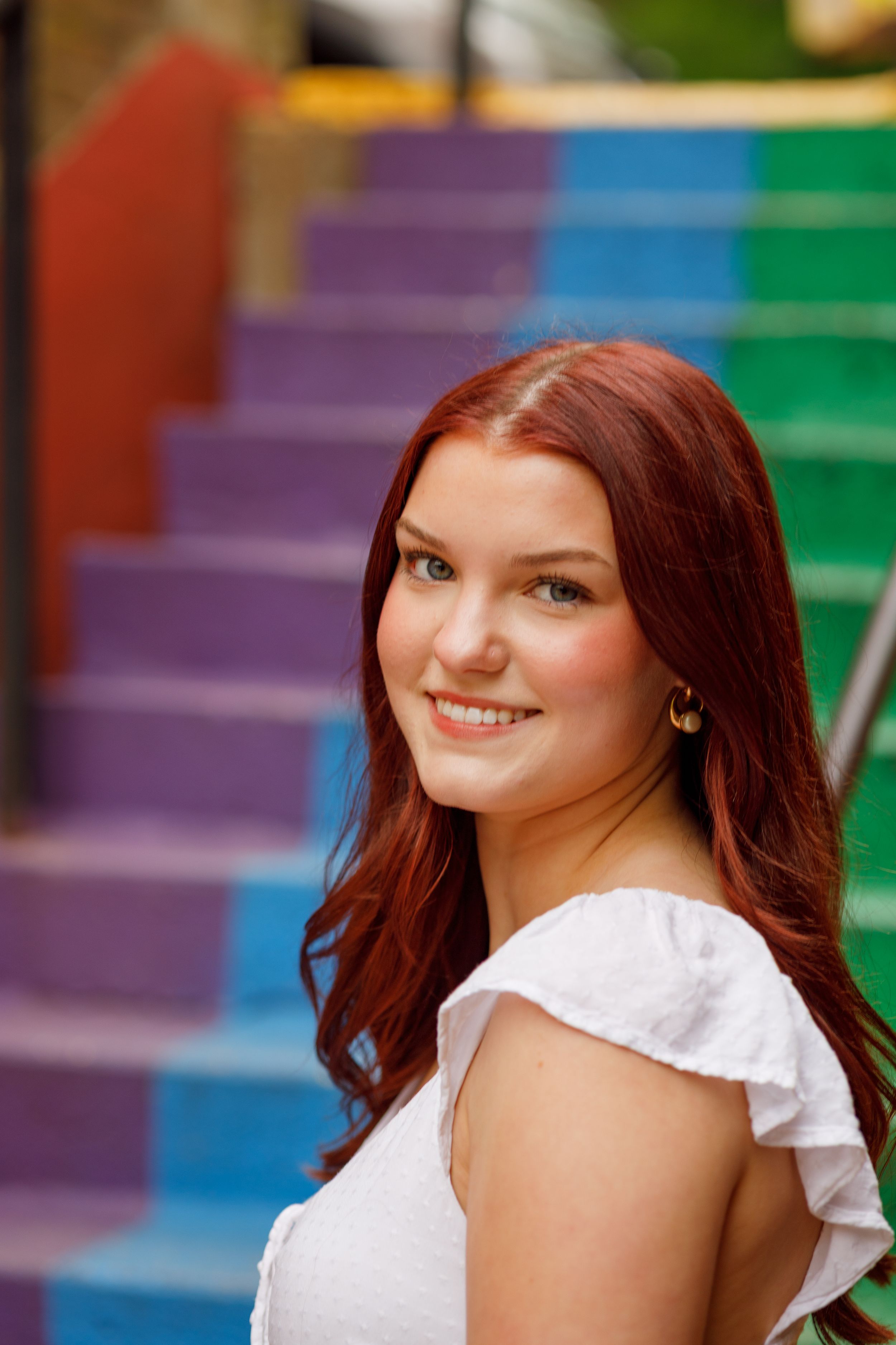 A woman in a white ruffled top poses on colorful rainbow painted stairs with vibrant red hair and a warm smile.