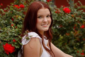Smiling woman in white summer dress poses in garden with vibrant red roses in background.