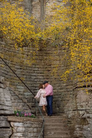 Two people share a romantic moment on stone steps surrounded by yellow blooming forsythia and an old stone wall.