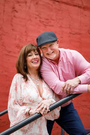 A couple in coordinating pink outfits share a joyful moment while leaning against a railing against a red wall.