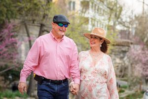 Smiling couple holds hands while walking in a spring garden with blooming cherry blossoms in the background.
