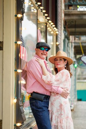 A couple in coordinating pink outfits poses together outside a storefront with string lights and an American flag.