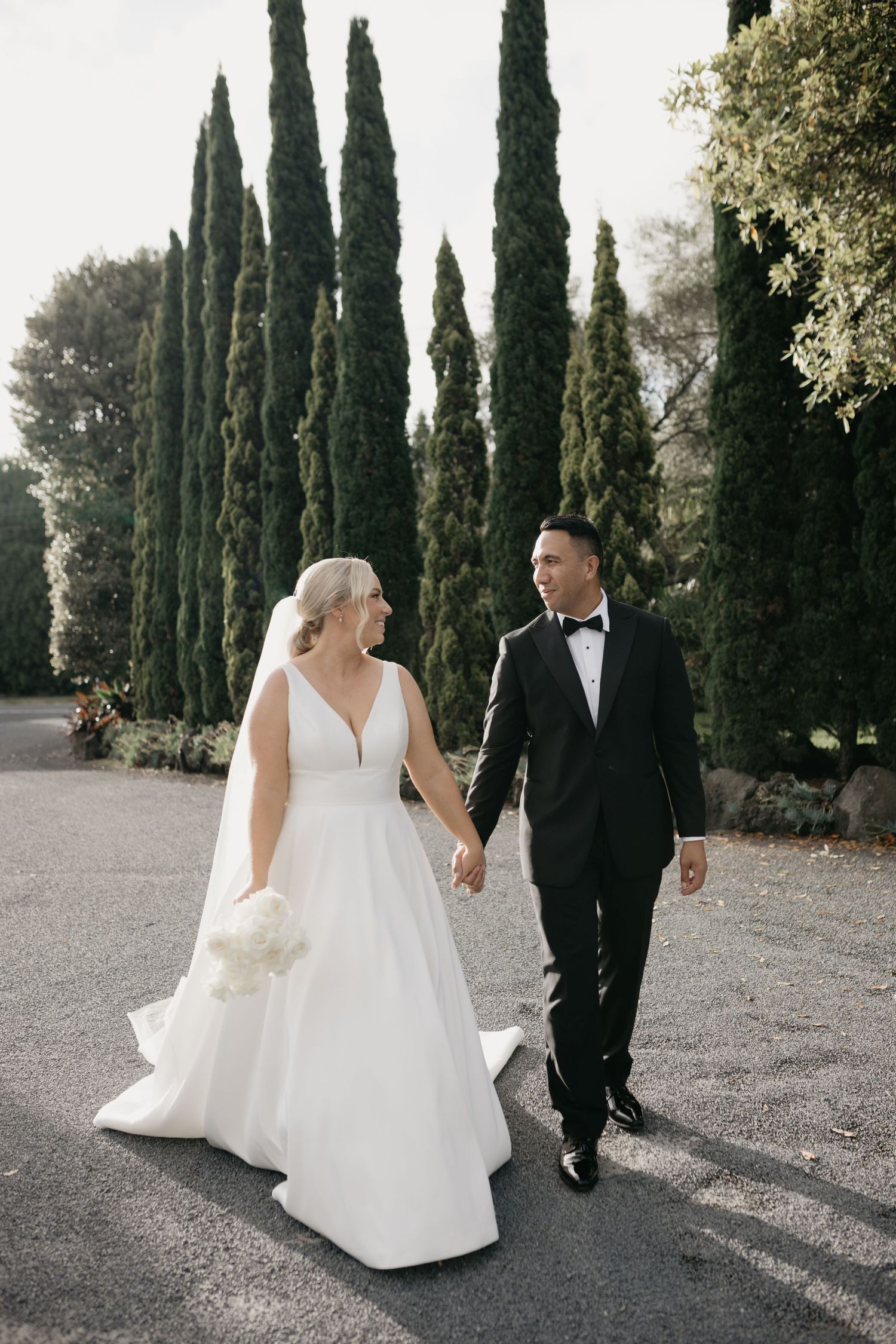 Bride in white gown and groom in tuxedo walking hand-in-hand past tall cypress trees.