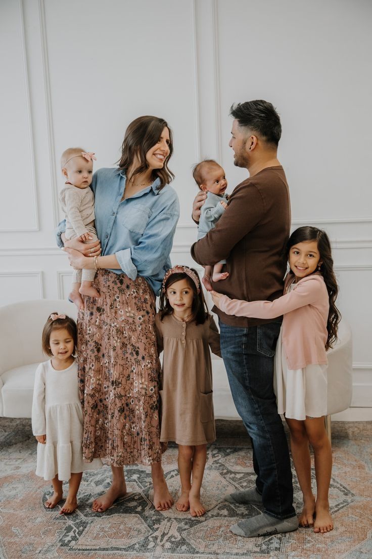 Family standing barefoot on patterned rug against white wall wearing coordinated casual outfits in neutral tones.