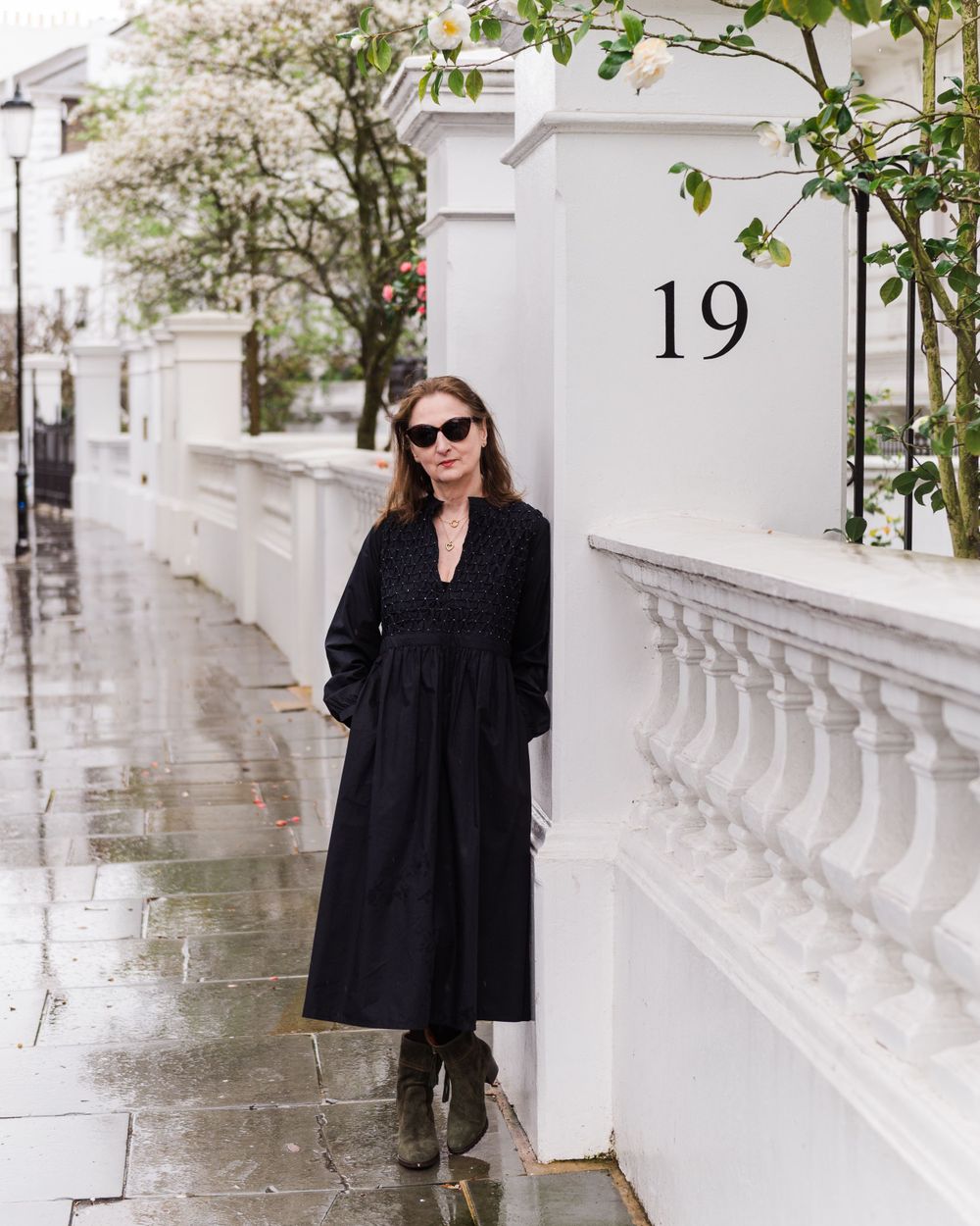 Person in a black dress and sunglasses poses against a white wall with climbing vines in an elegant outdoor setting.