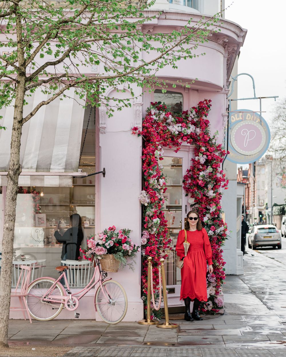 Pink storefront decorated with climbing red roses, pink bicycle, and figure in red dress standing by floral archway in Paris.
