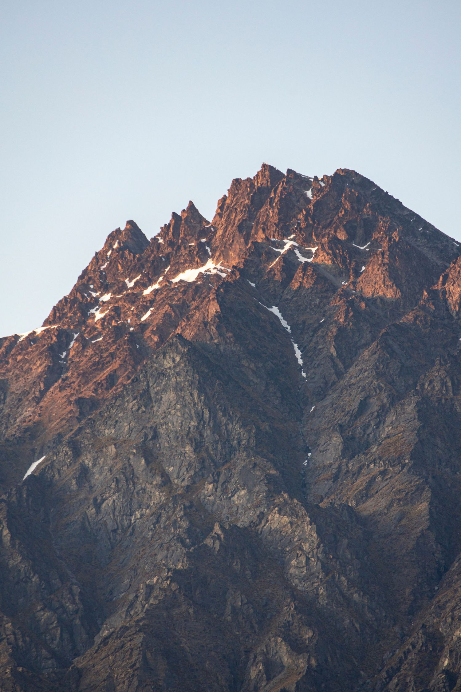 Rugged mountain peak with patches of snow, bathed in warm light at sunset against a clear blue sky.