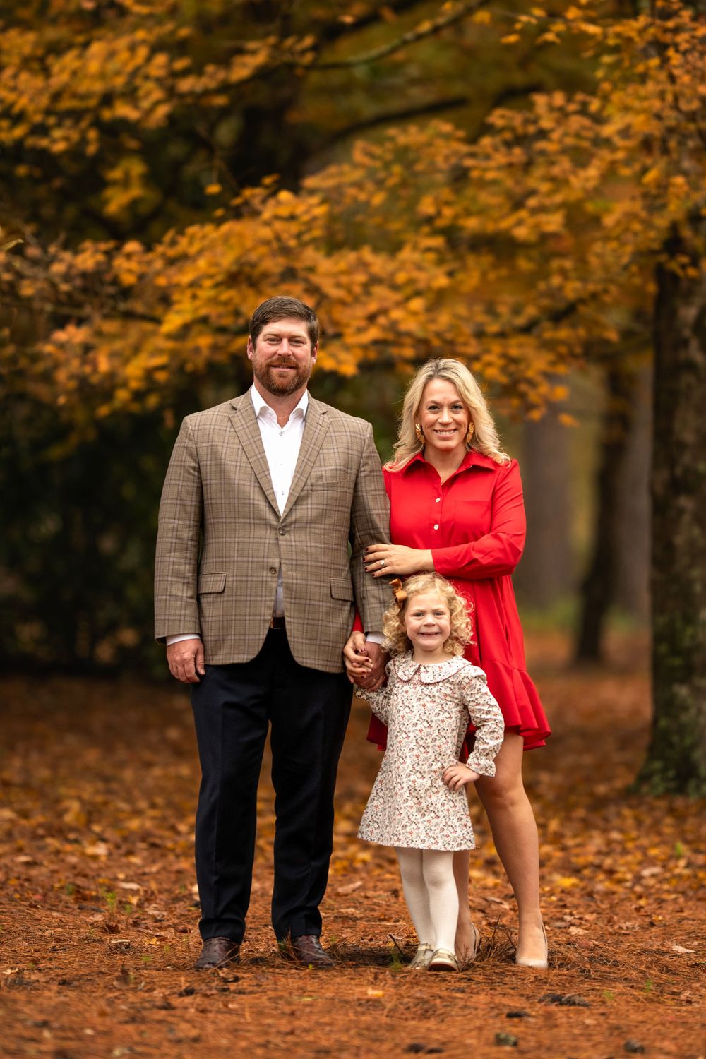 A cozy autumn family portrait in brown and red attire standing among fallen leaves in a wooded setting.
