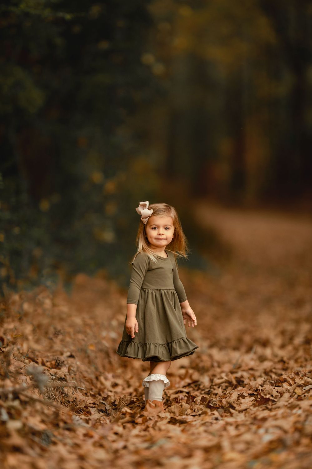 A small child in an olive dress plays in fallen autumn leaves.