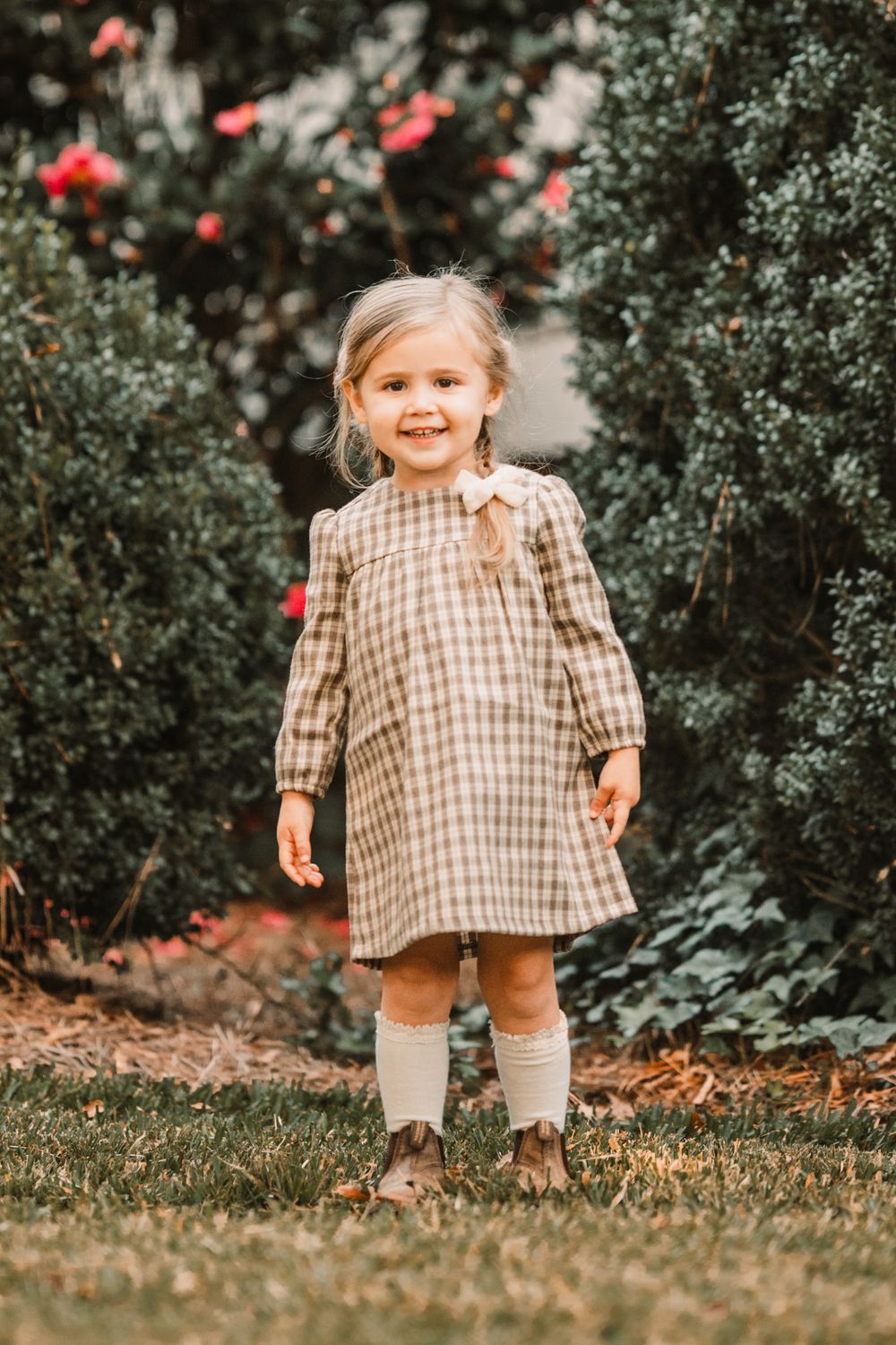 A child in a brown checkered dress plays outdoors among green foliage and red flowers.