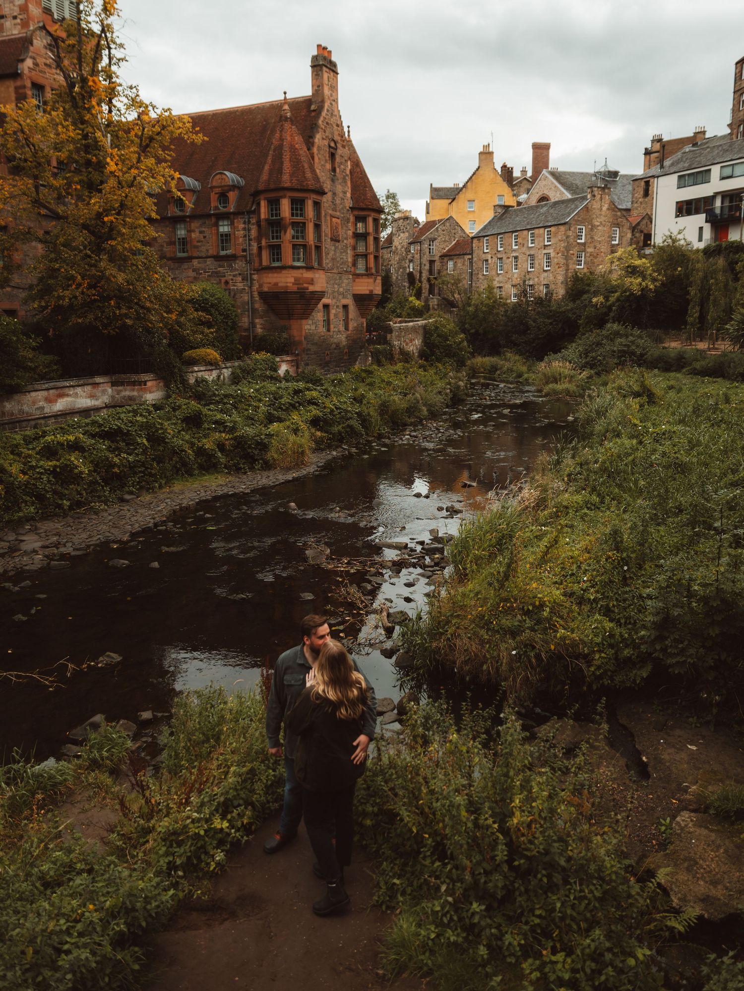 Autumn engagement photoshoot in a scenic location with historic buildings and water in the background.