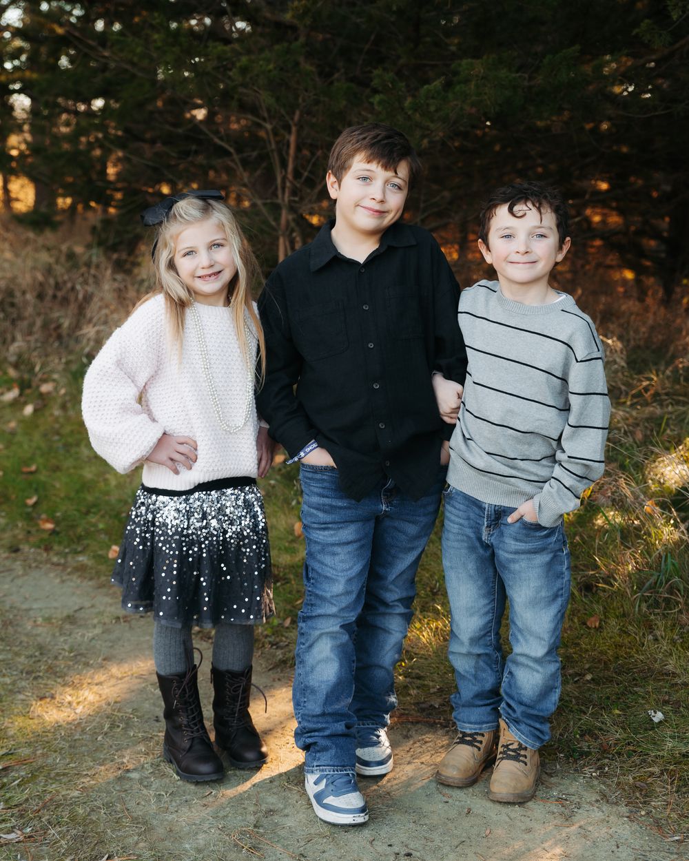 Three children standing together wearing casual clothes on a dirt path during sunset.
