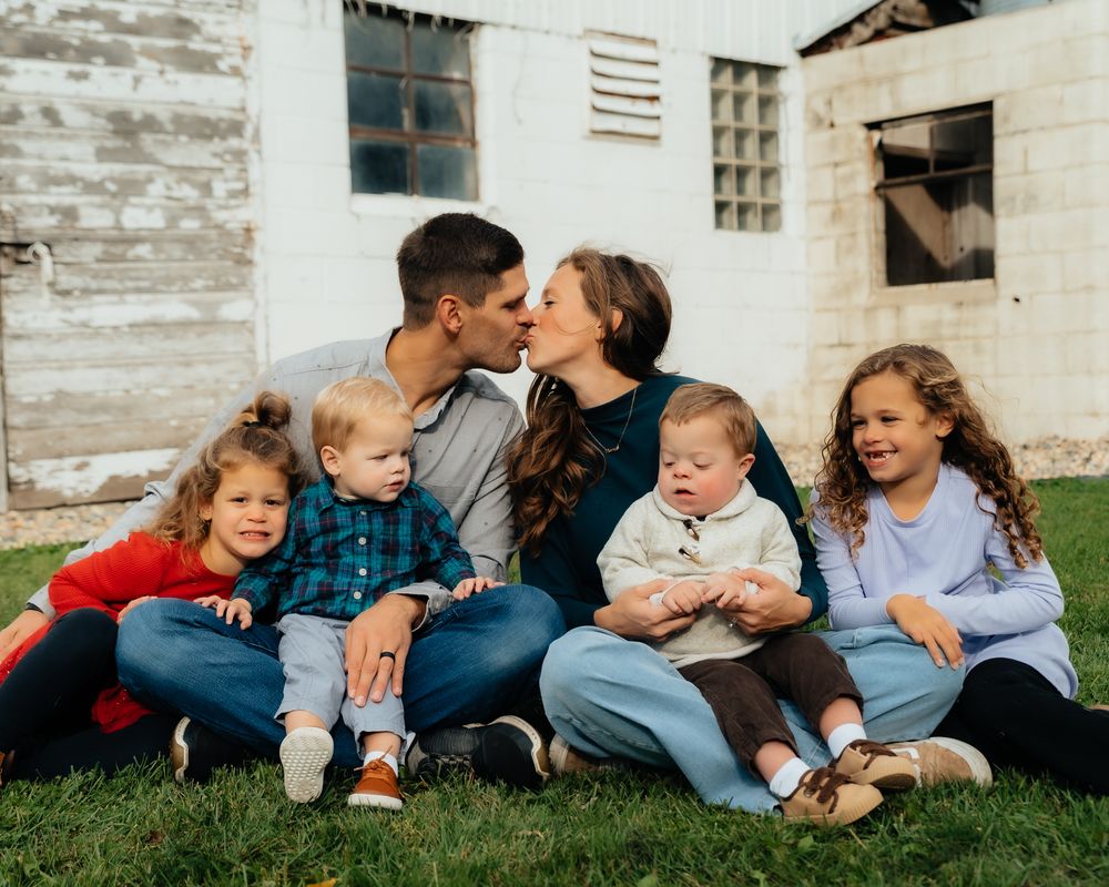 Family members relax on grass against a rustic white building backdrop during a photo shoot.