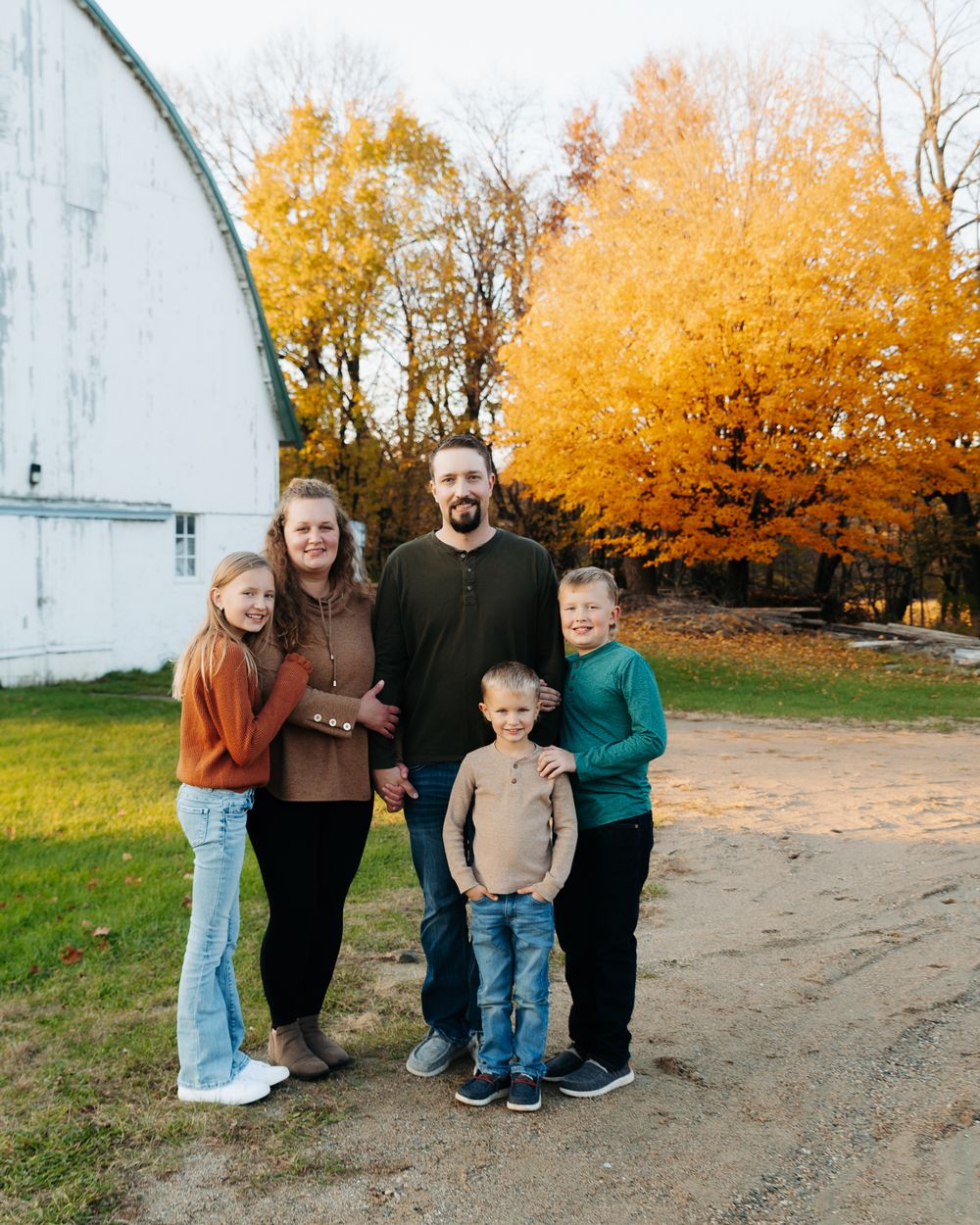 A group poses together on a dirt path with vibrant autumn trees and a white barn in the background.