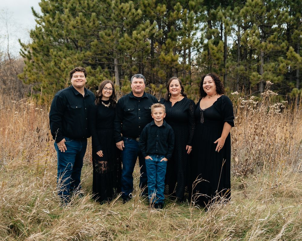 A panoramic outdoor family portrait session in a rustic field with tall grass and evergreen trees in the background.
