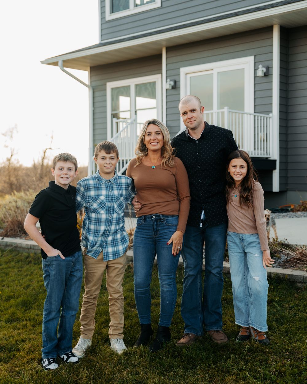 A family stands together outside their modern gray house on a grassy lawn at dusk.