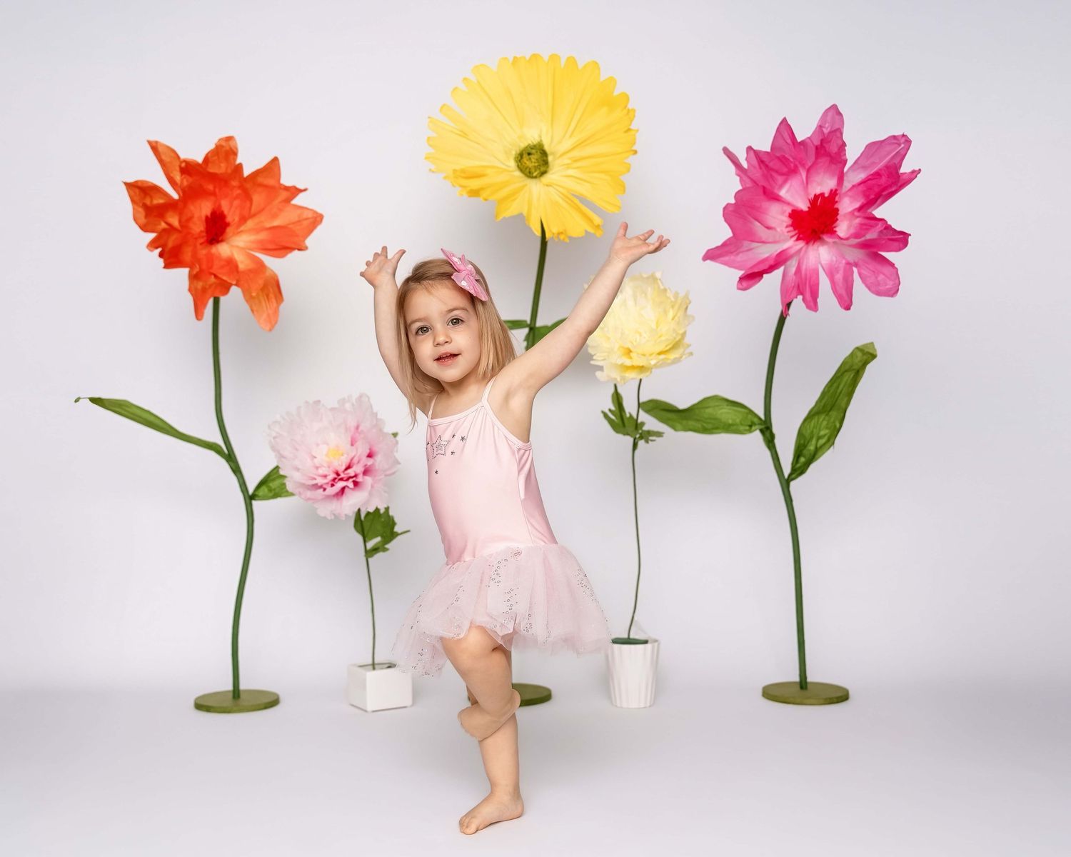 Studio portrait of a toddler dancing on white backdrop with giant flowers behind her, taken by Milwaukee family photographer Rachel Dickman.