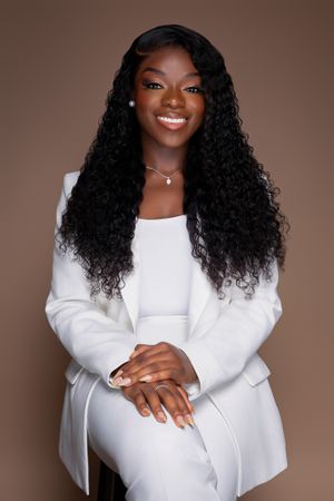 Professional portrait featuring elegant white blazer and long curly hair against brown background.