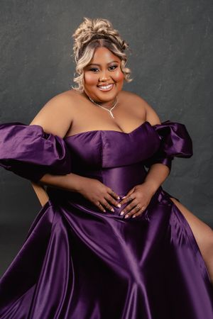 Woman in purple off-shoulder gown poses against dark background.