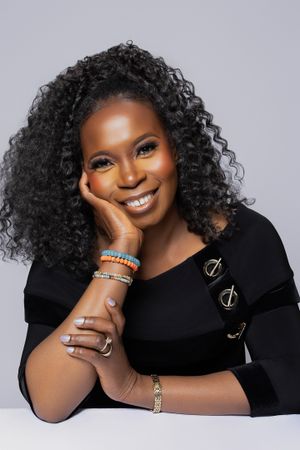 Professional headshot with natural curls, glowing makeup and black top against gray background.