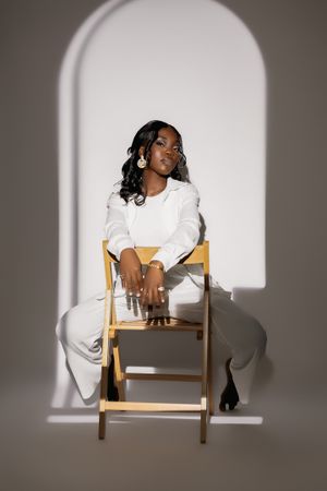 Stylish portrait in white attire sitting on wooden chair in arched window setting.