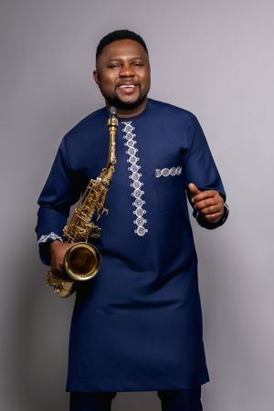 Musician in traditional navy blue African attire holding saxophone against grey background.