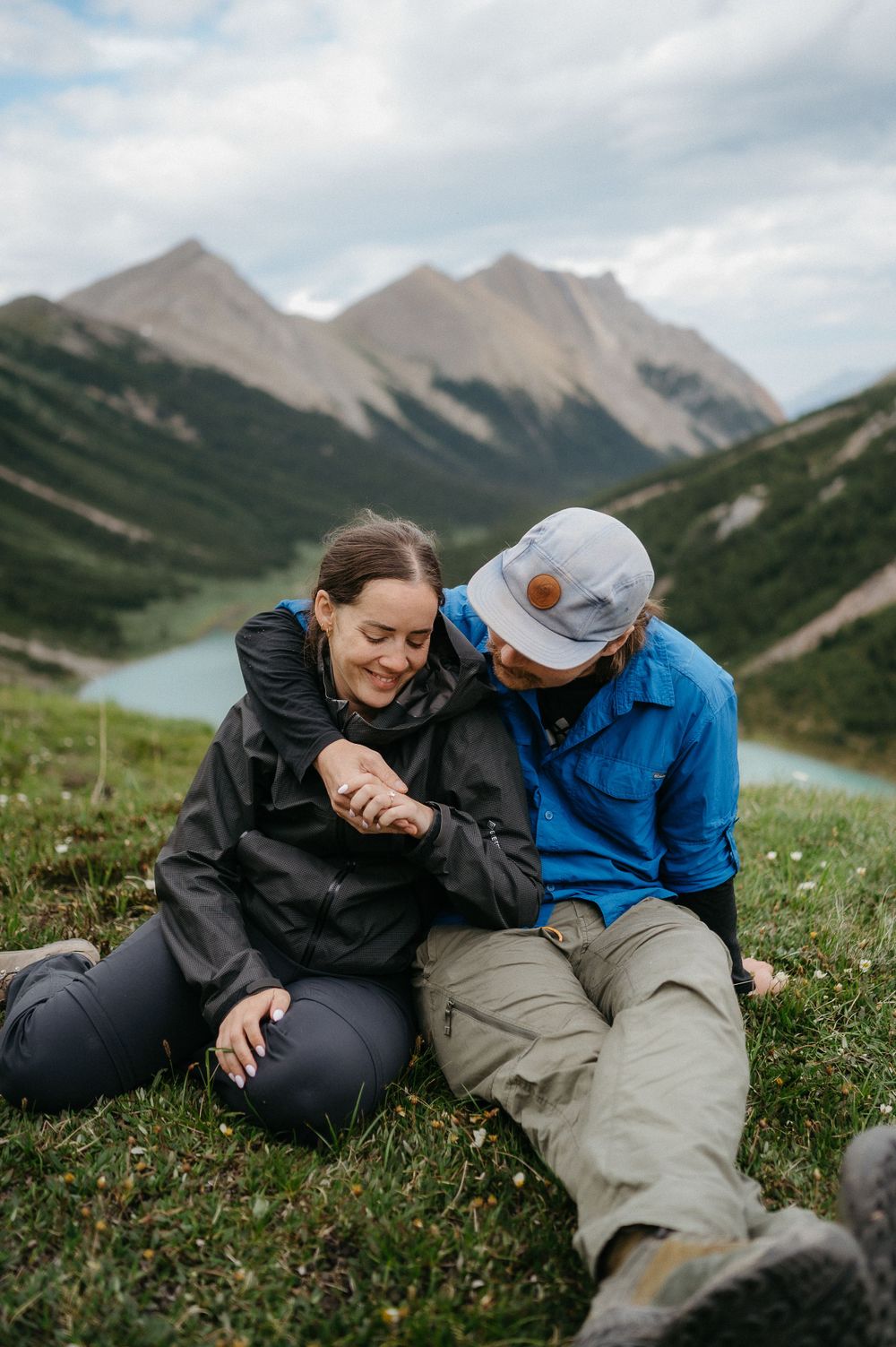 A couple shares a laugh while sitting in an alpine meadow with mountain peaks and a lake below.