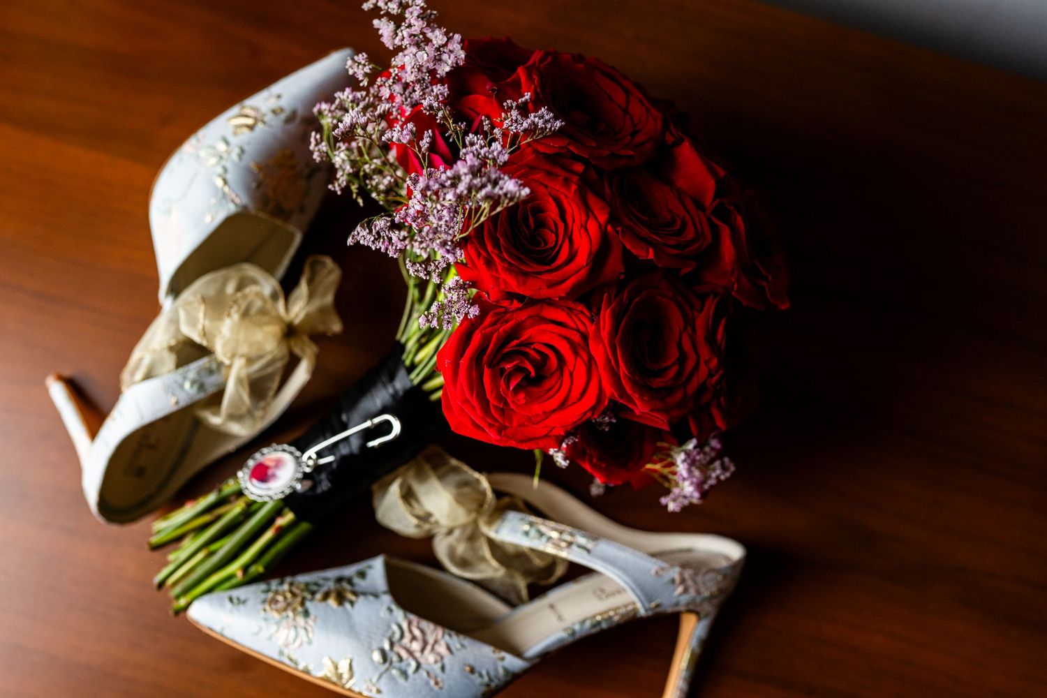A bouquet of red roses and purple flowers lays next to white bridal shoes on a wooden surface.