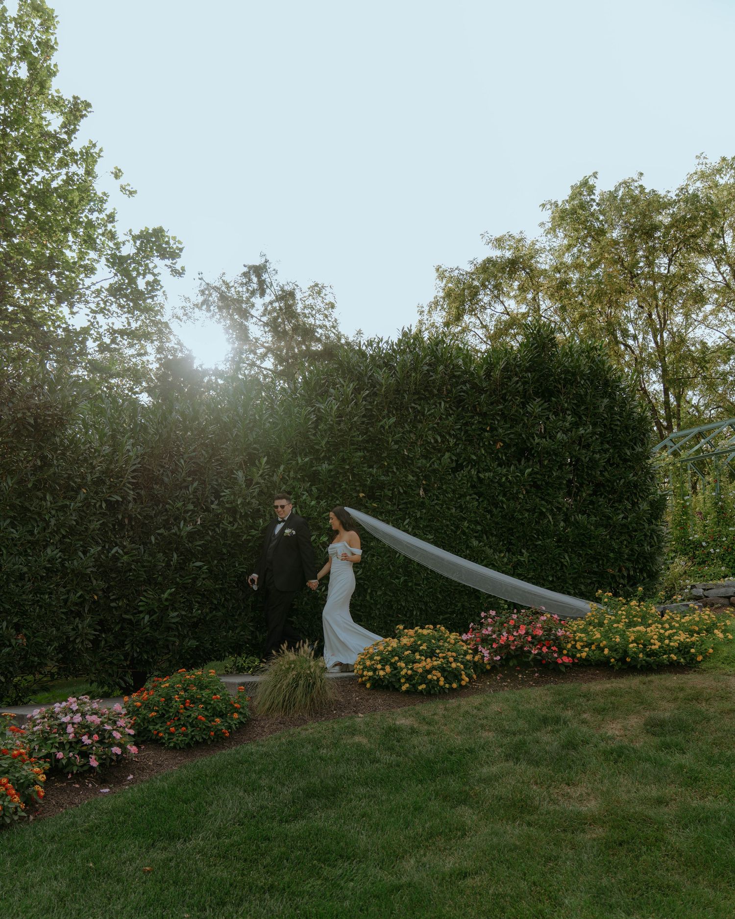 A bride in a flowing white gown with long train stands in a sunlit garden setting against stone walls and trees.