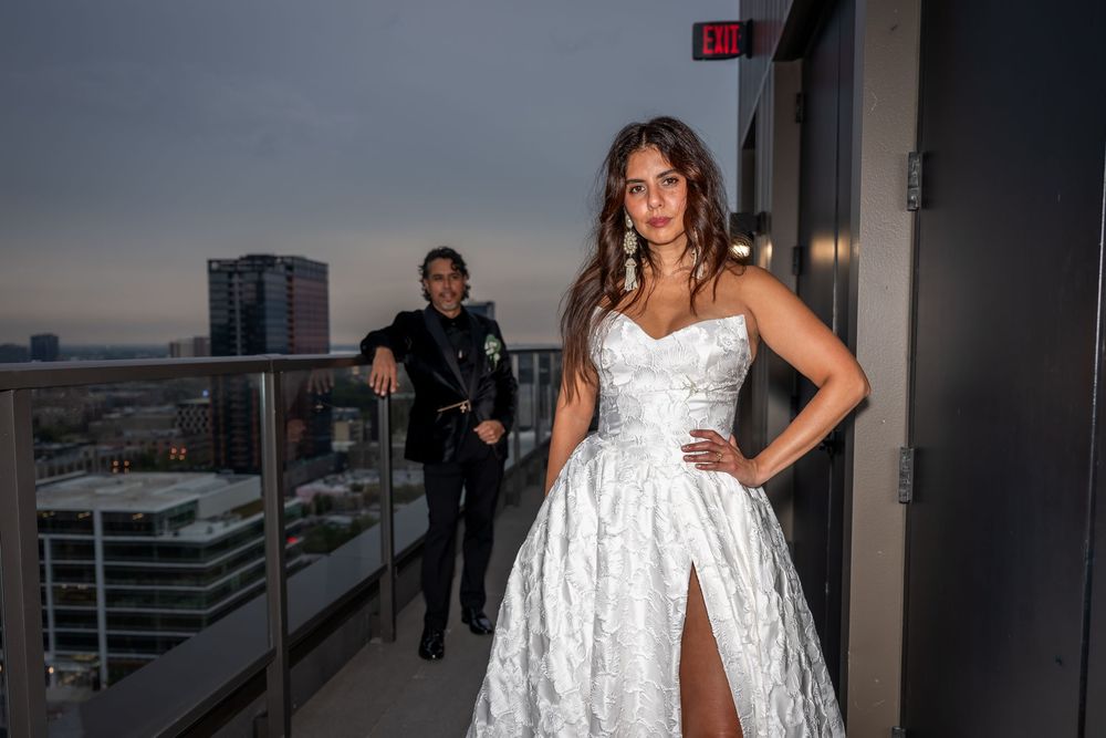 Bride in a white strapless gown stands confidently on a rooftop balcony at twilight with groom leaning on railing.
