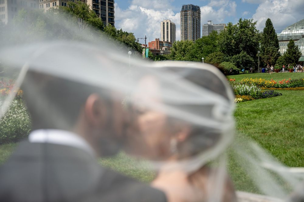 Bride and groom share a romantic kiss under a flowing veil in a lush green park with city skyline backdrop.