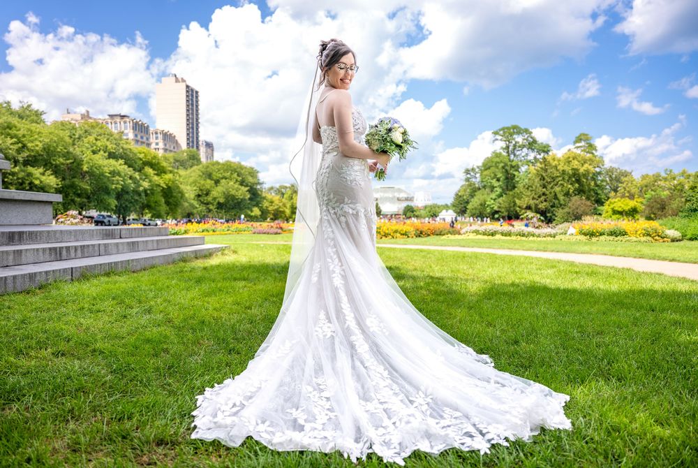 Bride in elegant lace mermaid wedding gown with long train poses in lush green park with city skyline backdrop.