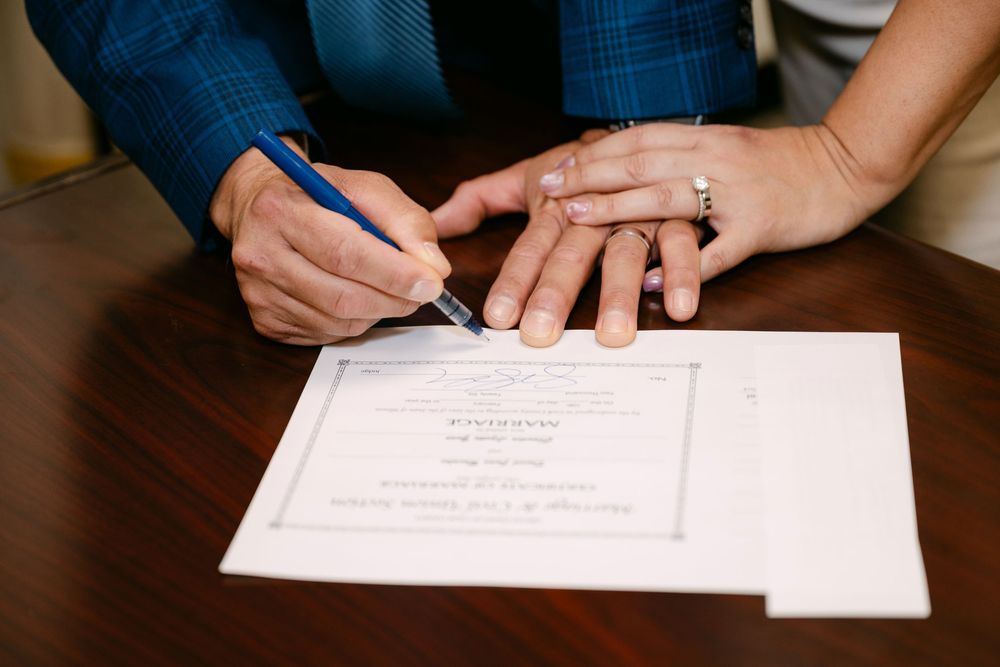Couple signing a marriage certificate at a wooden table, with engagement ring visible on bride's hand.