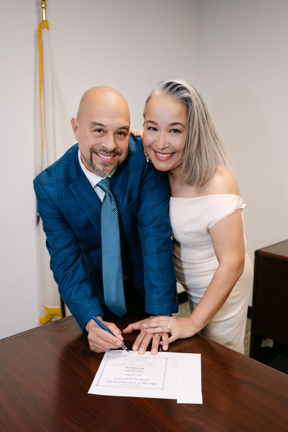Couple in wedding attire signing marriage certificate at a desk, smiling at camera in an official office setting.