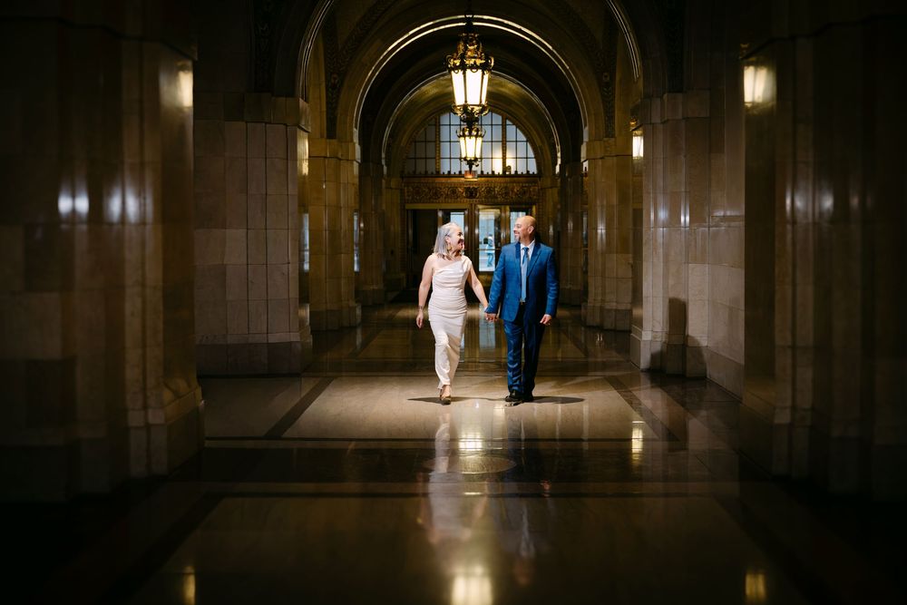 Elegant couple walking hand in hand through a grand illuminated hallway with arched ceilings and ornate stone architecture.