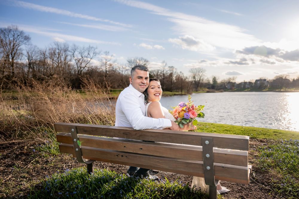Couple sitting on a park bench by a lake, woman holding colorful bouquet, smiling together on a sunny day.