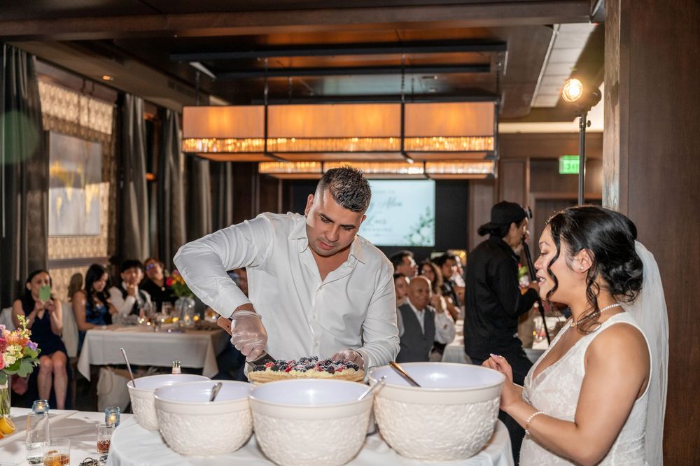 Bride and groom serving food from large bowls at wedding reception while guests watch in elegant venue.