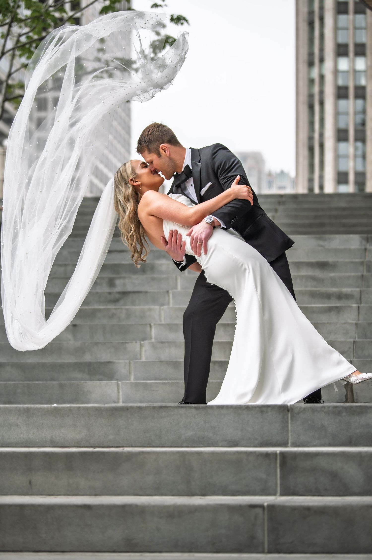 Groom dips bride on stone steps as her veil dramatically flows in the wind in the city.
