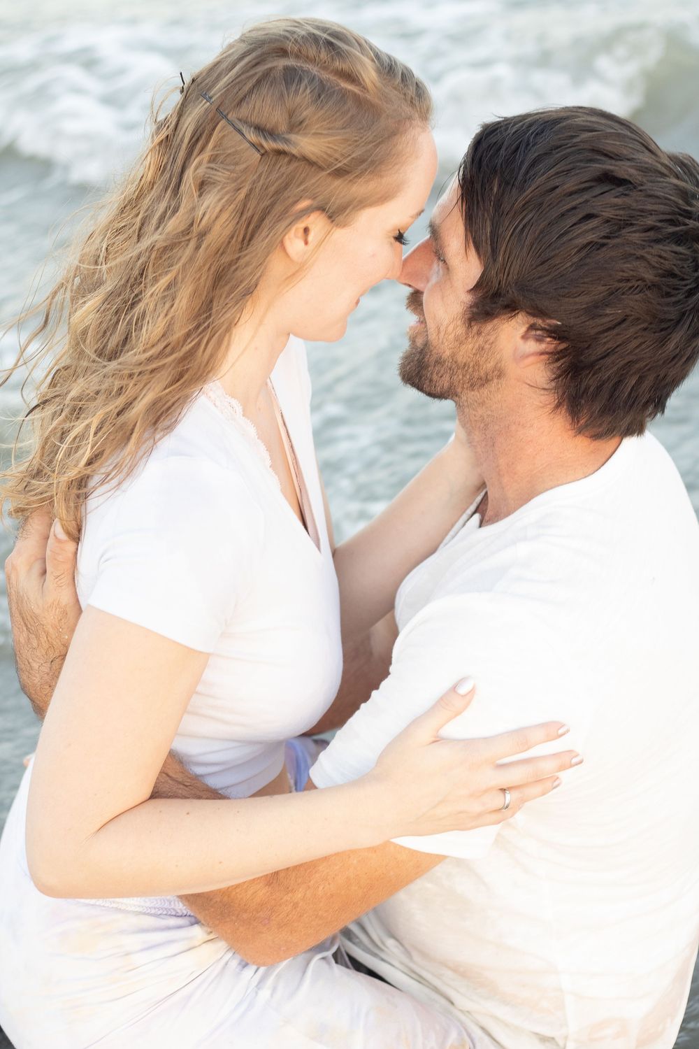 Romantic couple in white embracing on a beach, about to kiss with ocean waves in the background.