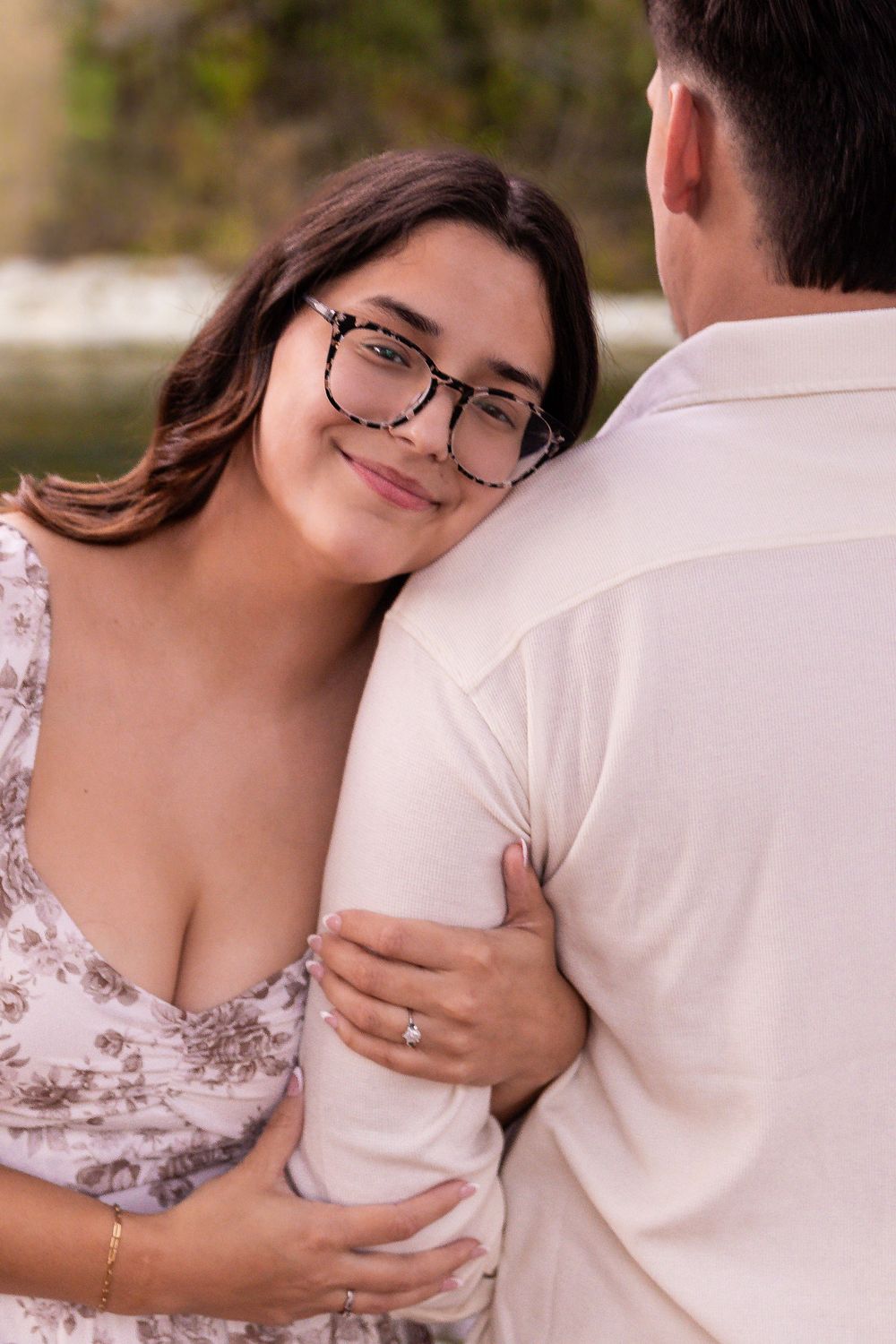 Woman with glasses in floral dress rests head on mans shoulder while showing off engagement ring outdoors.