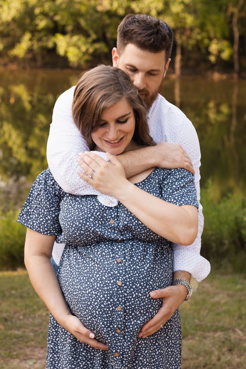 A man tenderly embraces a smiling pregnant woman in a blue floral dress outdoors near a serene pond.