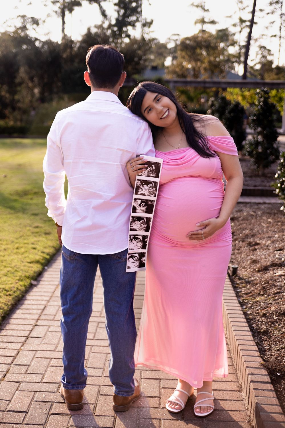 Pregnant woman in pink dress holds ultrasound photos with partner on brick path during outdoor maternity photoshoot.