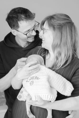 Black and white, first time parents, holding newborn baby in front of them. Baby is wearing a hand knitted cardigan with babies name on