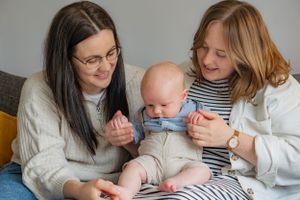 A natural family portrait of two mums with their baby boy. They are sat cuddling/tickling him, whilst looking at him and smiling.