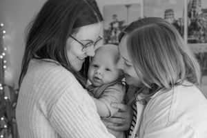 A black and white image of two mums having a cuddle with their baby boy. They are both looking at the baby with so much love.